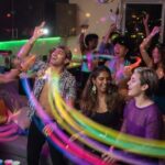 A vibrant, energetic party scene in a modern living room with diverse friends dancing together, colorful motion trails capturing movement, dynamic lighting reflecting off silhouettes, celebratory atmosphere, 16:9 wide cinematic shot, no text, no logos, no UI, no watermarks, safe margins 20%